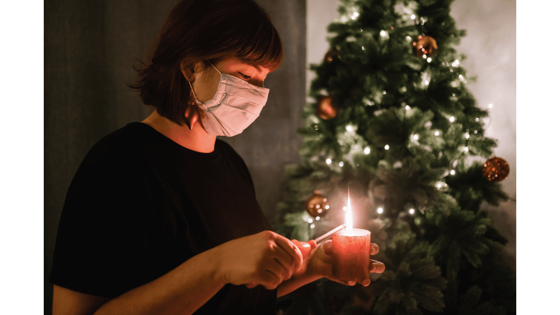 Woman in face mask lighting a candle in front of a Christmas tree