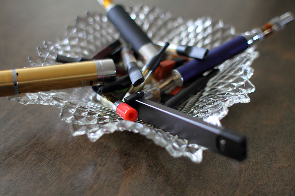 Closeup of assorted vape pens and cartridges in a glass ashtray.