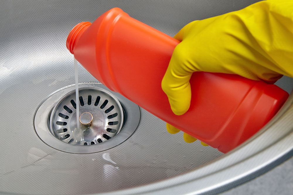 A hand with a yellow glove pouring drain cleaner into a sink
