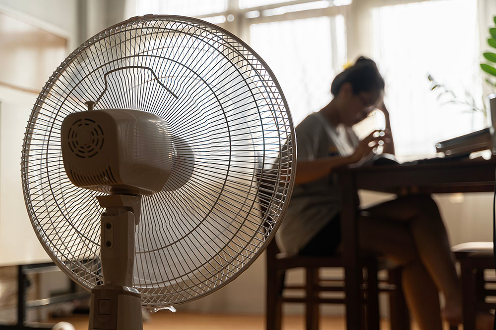 A woman sat in view of a standing fan to cool down